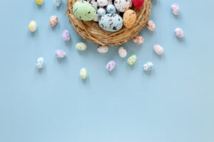 Niebieskie Tło z wiklinowym koszykiem z którego wysypują się kolorowe, pastelowe pisanki.