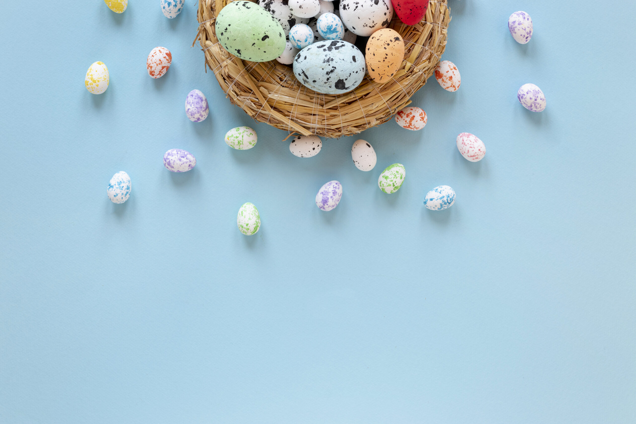 Niebieskie Tło z wiklinowym koszykiem z którego wysypują się kolorowe, pastelowe pisanki.